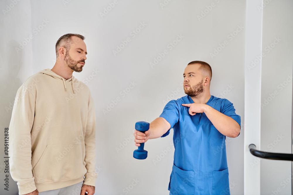 young doctor in blue uniform showing how to train with dumbbell to man in recovery kinesio center young doctor in blue uniform showing how to train with dumbbell to man in recovery kinesio center