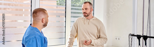 man talking to young rehabilitologist in blue uniform during appointment in kinesio center, banner photo