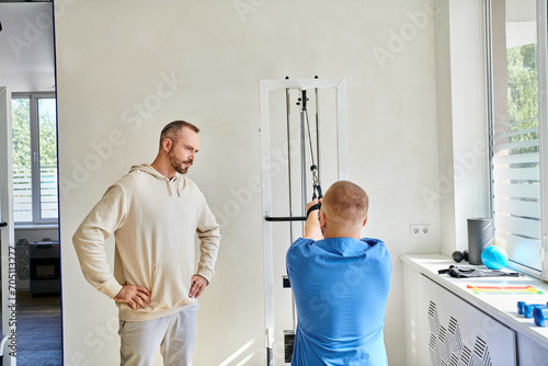 man looking at recovery expert in blue uniform training on exercise machine in kinesiology center
