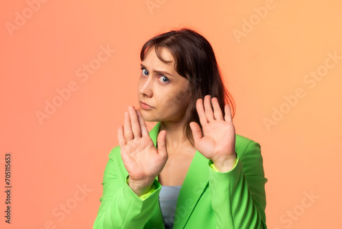 A woman in a light green suit on an orange background holds her hands in front of her, moving away from the interlocutor and as if saying, take it easy, I'm not ready for this, stay away from me.