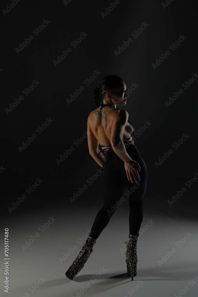 Young woman in black open clothes poses on black background of backlit studio. The dancer demonstrates elements of dance choreography in high heels. The concept of the dance promo shot.
