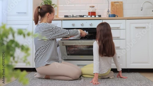 Smiling mother and daughter taking baking out of oven sitting near oven smelling delicious sweets kid helping mother on kitchen family posing at home while sitting on the floor. photo
