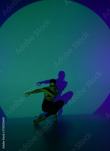 A young woman poses in a dark studio with blue light as the center of spotlight. The shadow of her body is reflected on the wall. A female dancer demonstrates elements of dance in high heels.