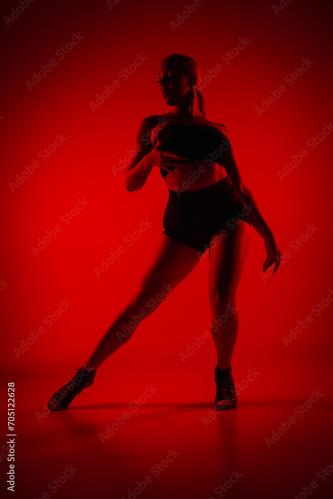 Silhouette female dancer in black shorts and top. Young woman poses gracefully and shows off slender flexible body in dark studio with red light. Elements of high heels dance choreography.