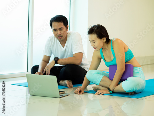 Two Asian young male and female adult sitting and looking vdo from laptop for practice Yoga pose demonstration in gym near window