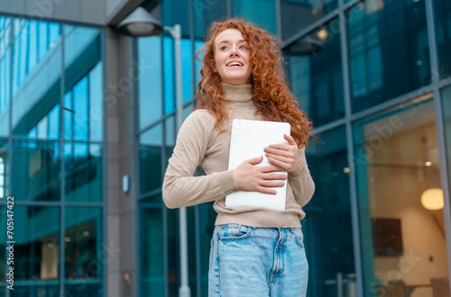 Portrait of beautiful happy young woman graduate with curly red hair holding laptop and excited  about news regarding her job application © Iryna