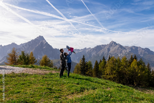 Happy family enjoying scenic view from lift station on mount Helm (Monte Elmo), Carnic Alps, Austria Italy border. Mother lifting up baby. Looking at mountains of untamed Sexten Dolomites South Tyrol photo