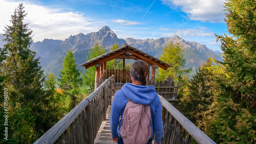 Hiker woman with backpack on wooden viewing platform with panoramic view of majestic mountain peaks of untamed Sexten Dolomites in South Tyrol, Italy, Europe. Hiking concept in Italian Alps in fall photo