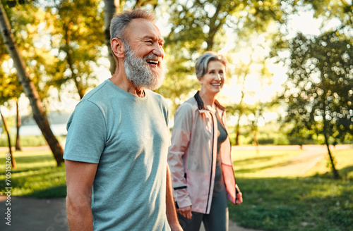 Active hobby on retirement. Grey haired caucasian man with beautiful wife strolling at summer park and smiling cheerfully. Fitness retired couple in sport attire taking break after morning run. photo