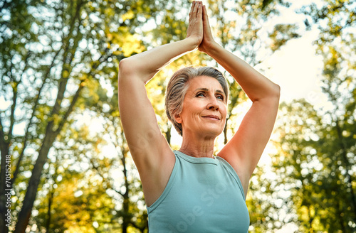 Spiritual meditation outdoors. Peaceful grey haired woman sitting at green sunny park with namaste hands over head. Caucasian lady in light blue tank top practicing yoga in harmony with nature.