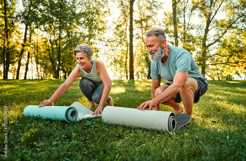 Outdoors fitness of couple. Sporty family of two old people with bare feet laying yoga mats on grass to exercise on fresh air. Retired man and woman preparing for joint yoga practice at sunny park.