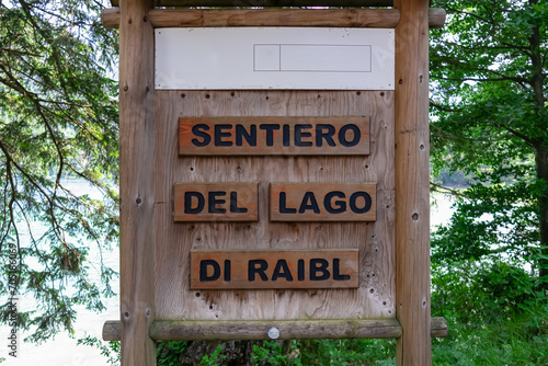 Informational sign of trailhead Sentiero Del Lago di Raibl at alpine lake Predil, Julian Alps, Tarvisio, Friuli Venezia Giulia, Italy. Signpost mounted on wooden post surrounded by trees and greenery