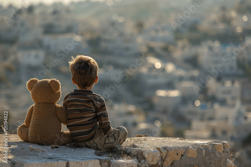 A little boy next to a teddy bear sits on the outskirts of the city and looks out over the ruined city after the disaster of the war earthquake