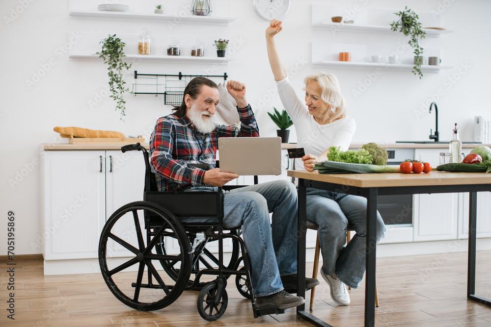 Happy Caucasian couple, pretty wife and bearded man in wheelchair, pay for food with credit card using laptop against background of spacious kitchen, happy with purchases. Happy Caucasian couple, pretty wife and bearded man in wheelchair, pay for food with credit card using laptop against background of spacious kitchen, happy with purchases.