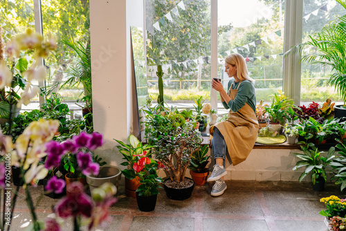 Happy small business female owner relaxing drinking coffee at flower shop sitting surrounded by plants at windowsill having break.
