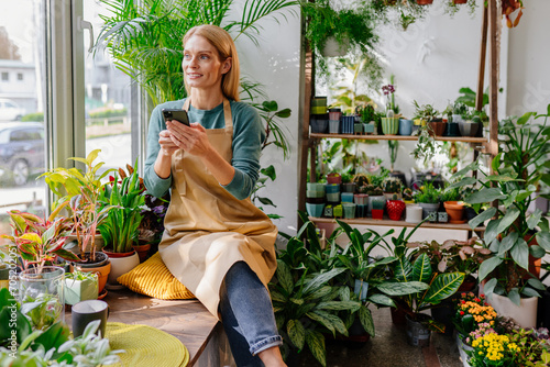 Beautiful smiling blonde middle age woman sitting with phone near window among flowers and browsing messages or websites in cell phone in interior of store with plants.