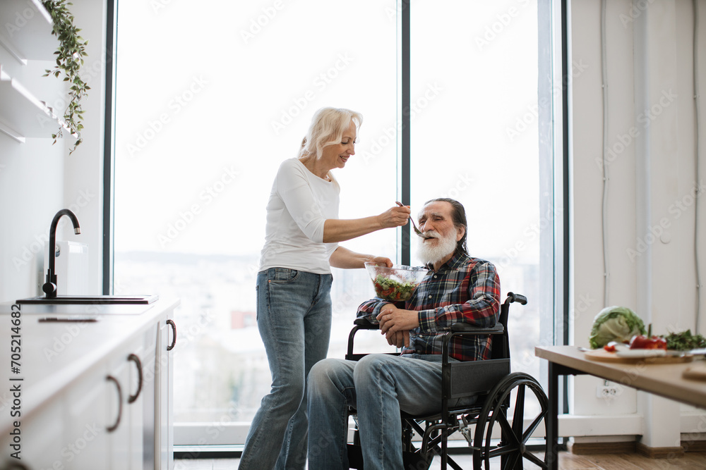 Happy elderly disabled couple having breakfast in modern kitchen with panoramic windows. Devoted woman feeds man in wheelchair with fresh healthy salad. Happy elderly disabled couple having breakfast in modern kitchen with panoramic windows. Devoted woman feeds man in wheelchair with fresh healthy salad.