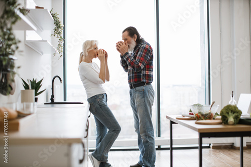 Happy elderly pensioners blond wife and husband with gray beard drinking hot drinks from mugs after delicious breakfast in bright modern spacious kitchen with panoramic windows.