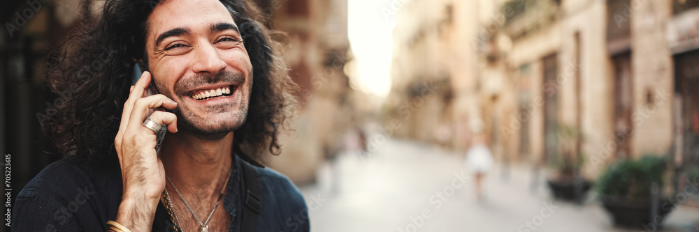 Young attractive italian guy with long curly hair and stubble is using mobile phone at old buildings background. Stylish man with an earring in his ear and lot of chains emotionally talking on phone - obrazy, fototapety, plakaty 