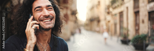 Young attractive italian guy with long curly hair and stubble is using mobile phone at old buildings background. Stylish man with an earring in his ear and lot of chains emotionally talking on phone photo