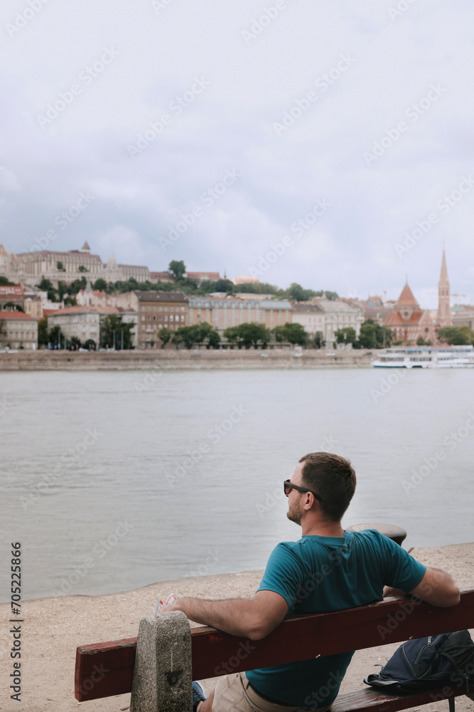 A young man sits on a bench on the Danube River embankment and looks at the panorama of the city of Budapest