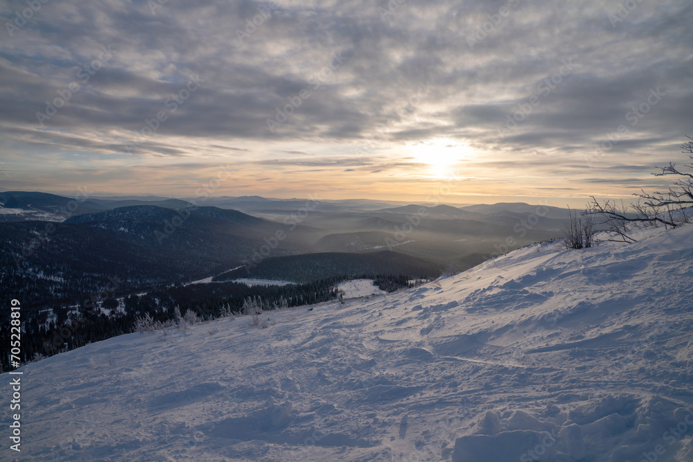 View from the top of the ski slope against the backdrop of mountains and forest under the evening sky.