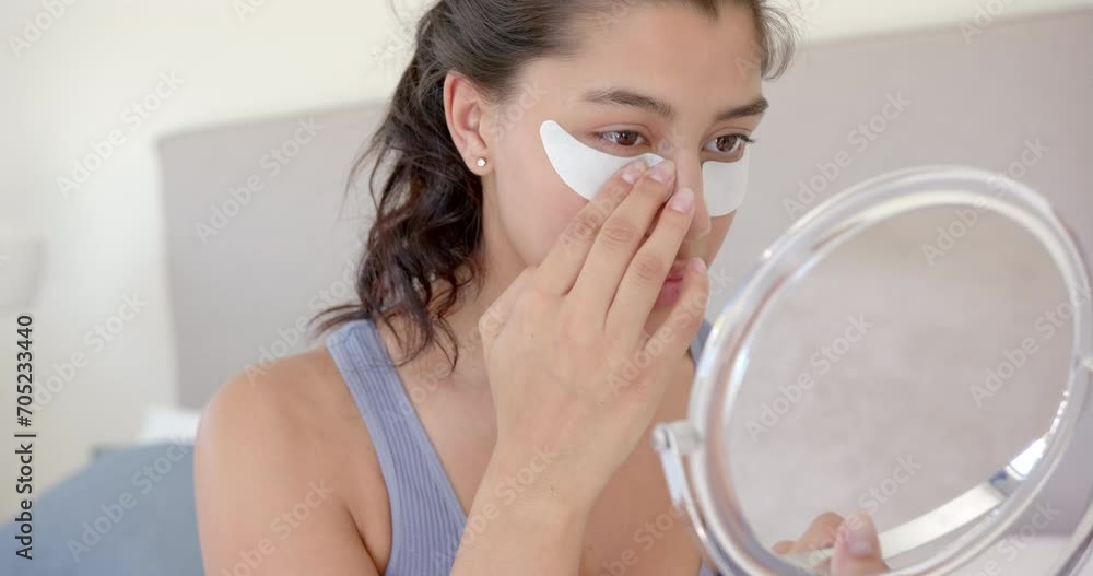 Focused biracial teenage girl putting on under eye masks looking in handheld mirror, slow motion Focused biracial teenage girl putting on under eye masks looking in handheld mirror, slow motion