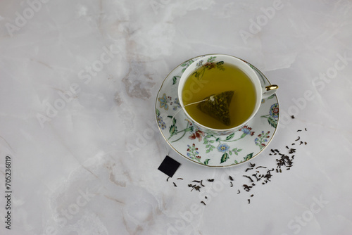 Bag of green tea with water in a porcelain cup and saucer with a picture of color stands on a white marble table