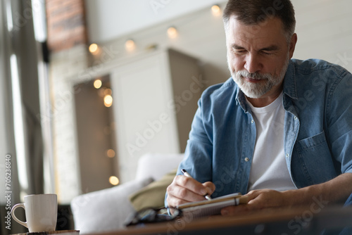 Senior man taking notes in notebook while sitting on the couch, gray-haired elderly man writing thoughts in notebook photo