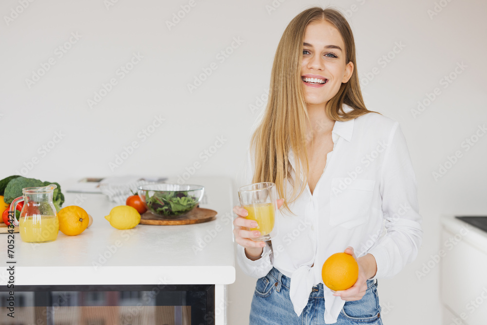 A happy cute blonde woman drinks orange juice in the kitchen - obrazy, fototapety, plakaty 