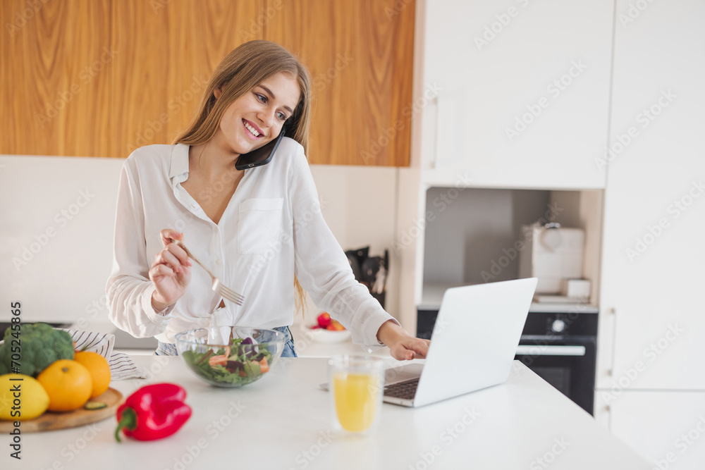 Happy and smiling blonde woman talking on the phone and looking for a recipe on the internet on a laptop in the kitchen Happy and smiling blonde woman talking on the phone and looking for a recipe on the internet on a laptop in the kitchen