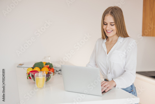 Happy and smiling blonde woman looking for recipe on laptop on internet in kitchen