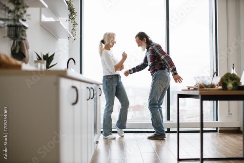 Caucasian old couple spends free time listening to music and dancing the twist in modern light kitchen. Seniors husband and wife having fun at the weekend in a cozy house.