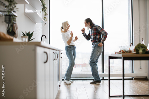 Caucasian old couple spends free time listening to music and dancing the twist in modern light kitchen. Seniors husband and wife having fun at the weekend in a cozy house. photo