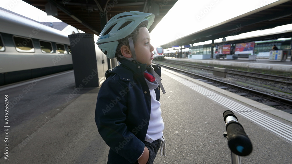Obraz premium One pensive young boy standing at train platform wearing helmet staring blankly into the horizon daydreaming. Thoughtful Child with hands in pocket waiting for transportation to arrive