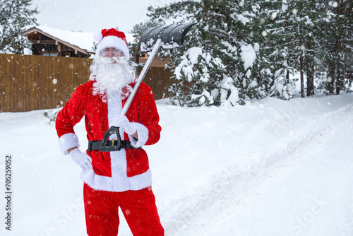 Santa Claus cleans snow with shovel in winter outdoors after a snowfall. Cleaning the streets in the village, clearing the passage for cars, difficult weather conditions for Christmas and New Year © Ольга Симонова