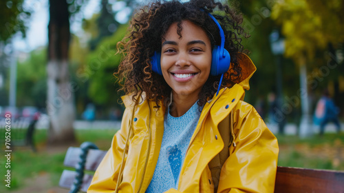 Young African American woman with a phone and blue headphones listening to music outdoors. A curly dark-skinned woman enjoys the weather while sitting on a bench in the park.