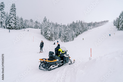 Man on a snowmobile rides past skiers descending a snowy slope. Back view