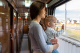 Mom holding her infant baby girl in her arms while riding in a public transportation. Cute toddler girl traveling with his mother. Family traveling in a train and looking through window.