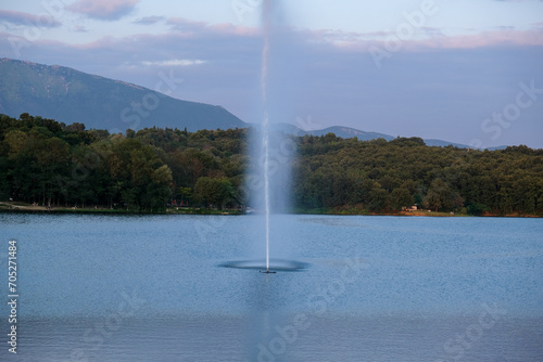 Tirana lake with a waterjet and breeze from a fountain, hills and green leafy trees on misty background.