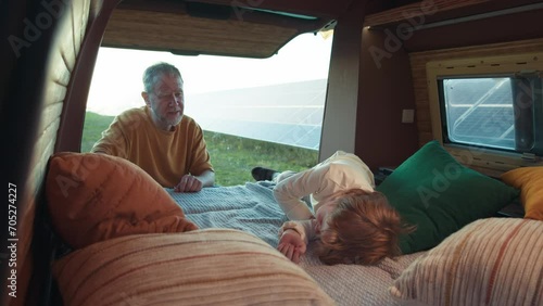 Inside the RV, a young boy lies sleeping on a bed with pillows, and grandfather sits beside him. The van is parked at a solar power plant, with photovoltaic panels visible through the window photo