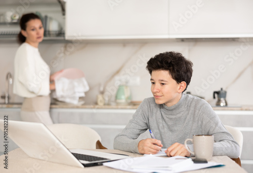 Boy studiyng using laptop, mother washing dishes and helping
