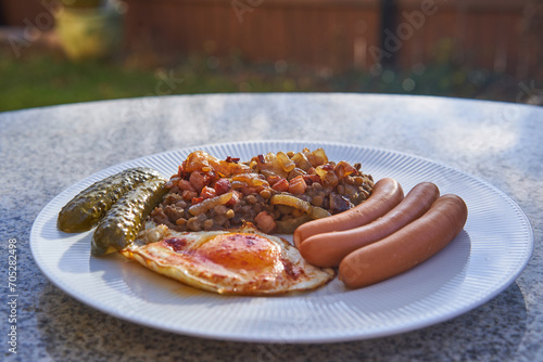 Traditional but delicious recipe from Czech cuisine, pork sausages and fried egg served with sweet and sour lentils mash and pickled cucumber outside on the stone table in the garden in sunny day.