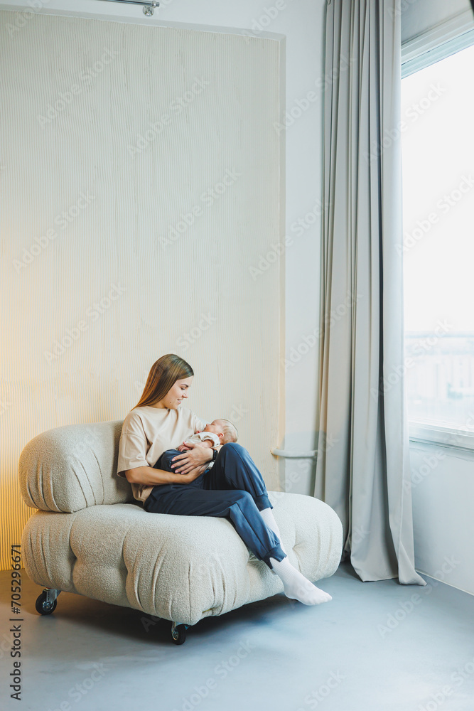 A smiling young mother is sitting on a sofa and holding a newborn baby in her arms. A happy mother with a child in her arms. The concept of motherhood