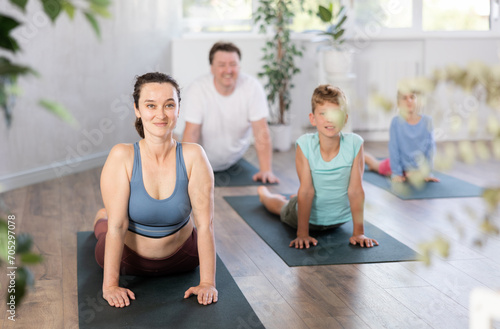 Diligent middle-aged woman practicing cobra pose of yoga on black mat together with her husband and children