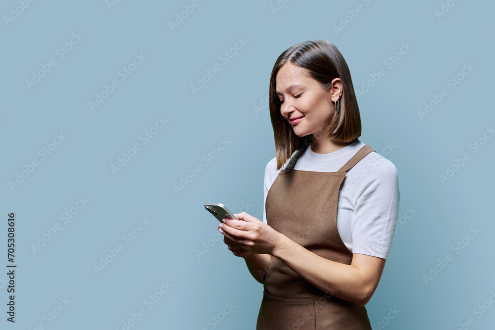 Portrait of young woman in apron holding smartphone in hands on blue background Portrait of young woman in apron holding smartphone in hands on blue background