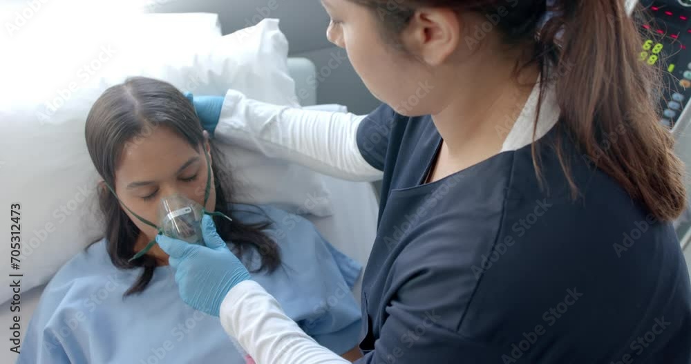 Diverse female doctor putting oxygen mask on female patient in hospital bed, slow motion