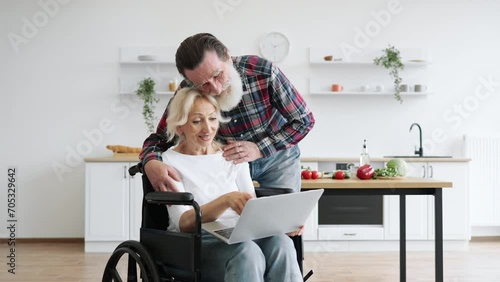 Close up of carming old couple of woman in wheelchair with laptop and husband hugging her on background of bright modern kitchen, looking at camera. photo