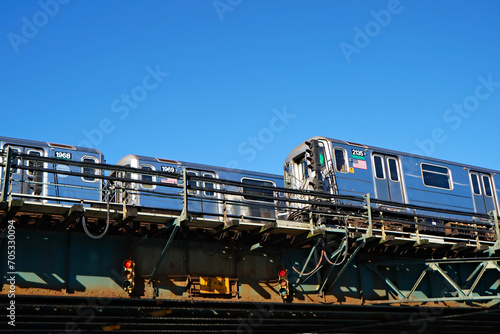 Two shiny bright metal silver elevated subway trains crossing under a clear blue sunny sky.