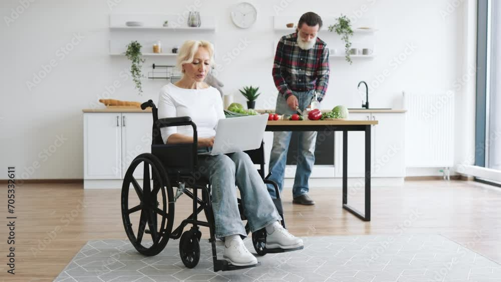 Full length portrait of old woman sits in wheelchair and works on laptop while her husband with gray beard prepares healthy salad in kitchen in background. Full length portrait of old woman sits in wheelchair and works on laptop while her husband with gray beard prepares healthy salad in kitchen in background.